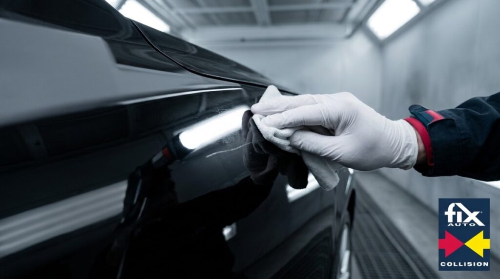 Gloved hand wiping a scratch on a black car door, showing clear coat damage in a brightly lit garage.
